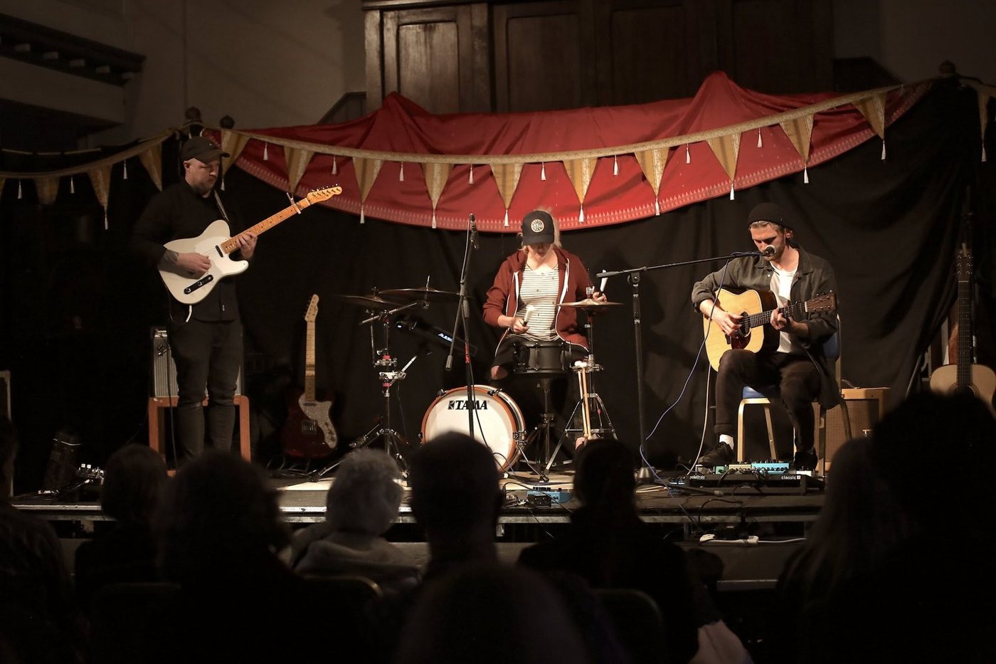 The Ollie Dixon Trio performing beneath red bunting with an audience at Ashburton Arts Centre