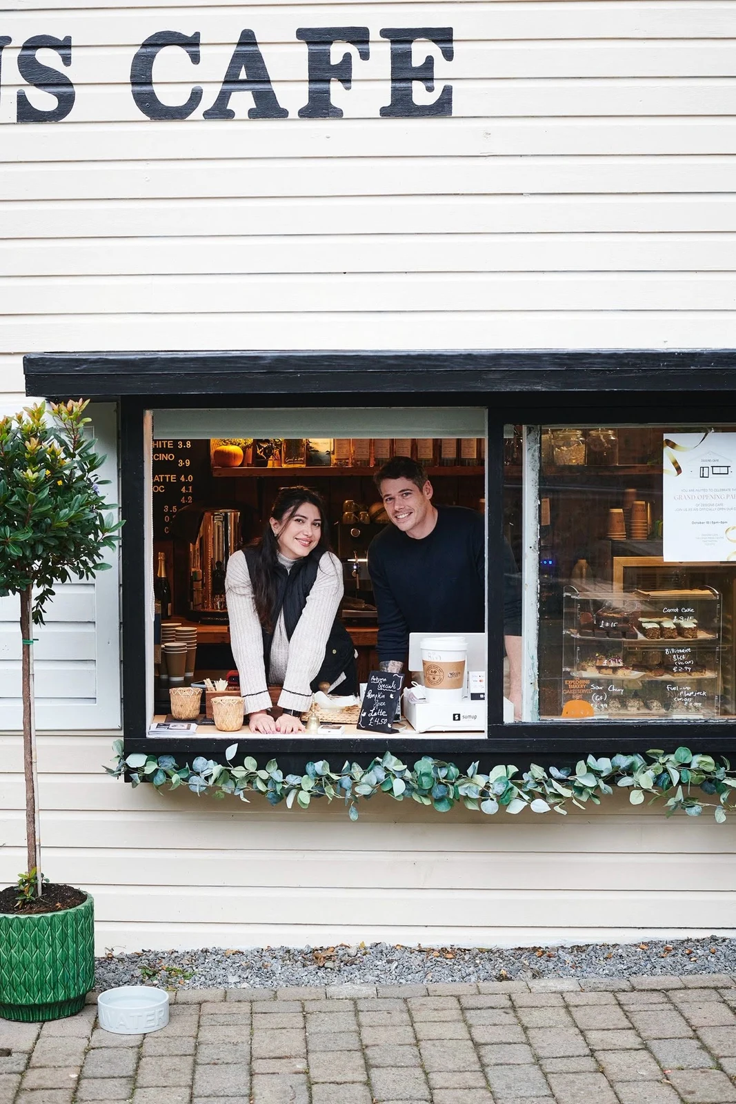 Two people at the Designs Café serving window with coffee and cakes on display