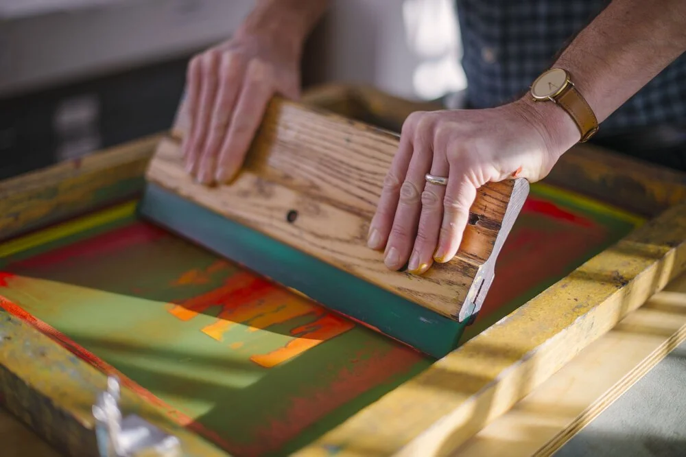 Hands pulling a screenprinting squeegee across a brightly coloured screen