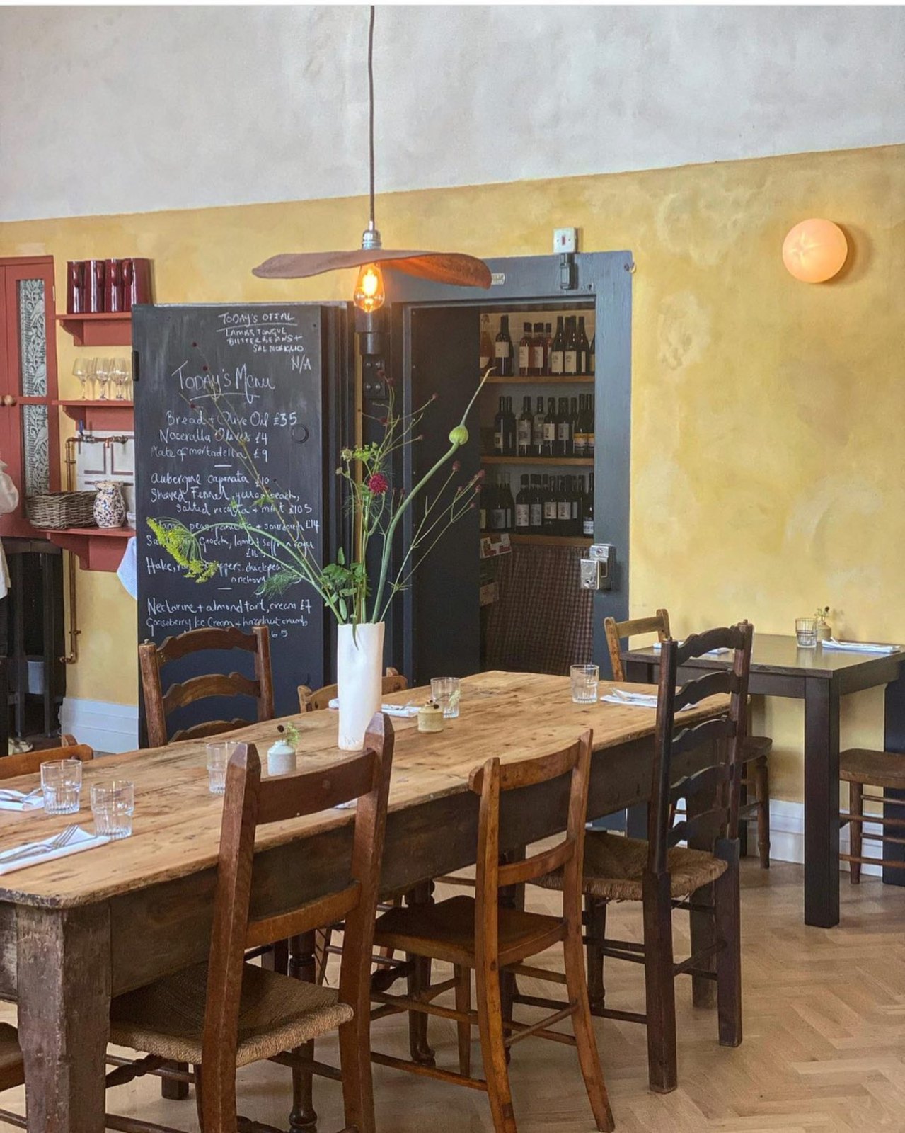Emilia’s long wooden dining table with flowers, warm yellow walls, a handwritten menu and wine bottles in the old vault
