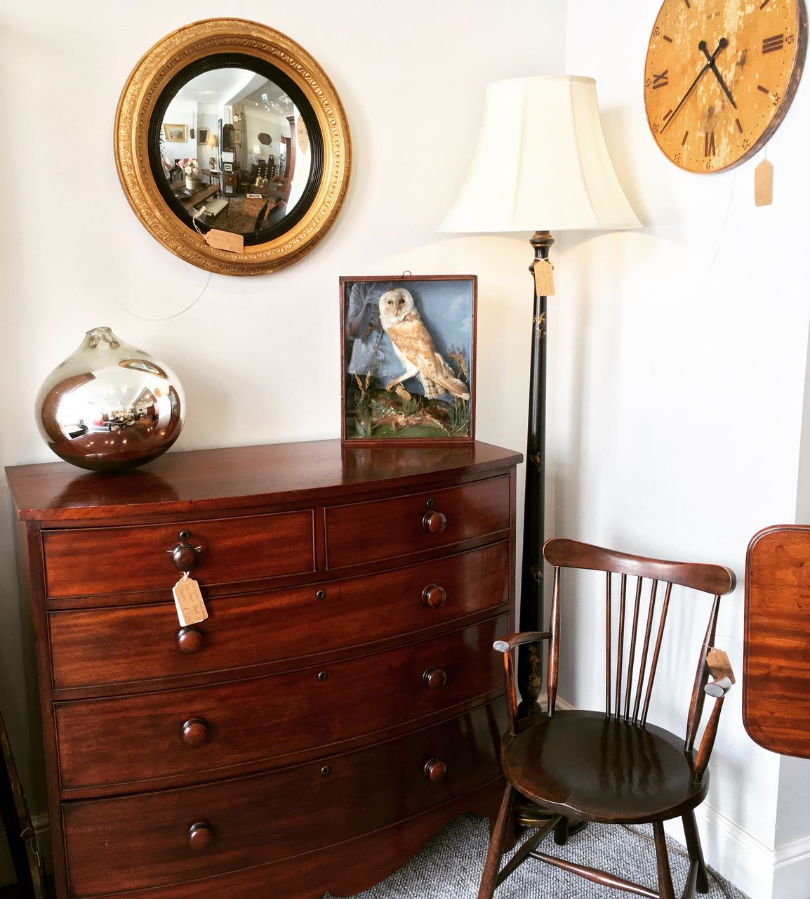 An antique chest of drawers beneath a round mirror, with lamps and a chair