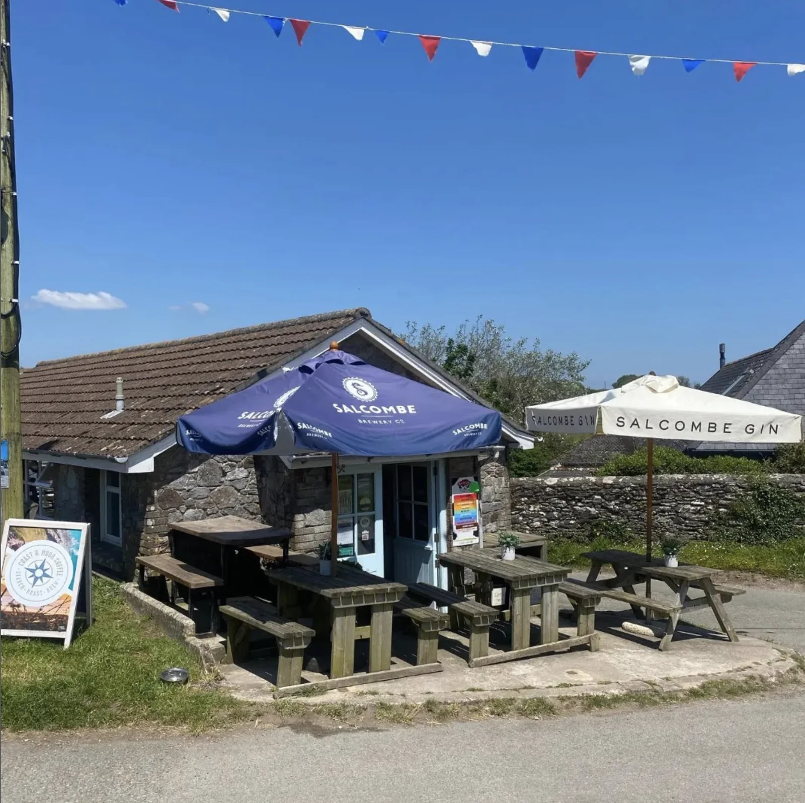 Stone café with picnic tables, blue parasols and bunting beside the village green