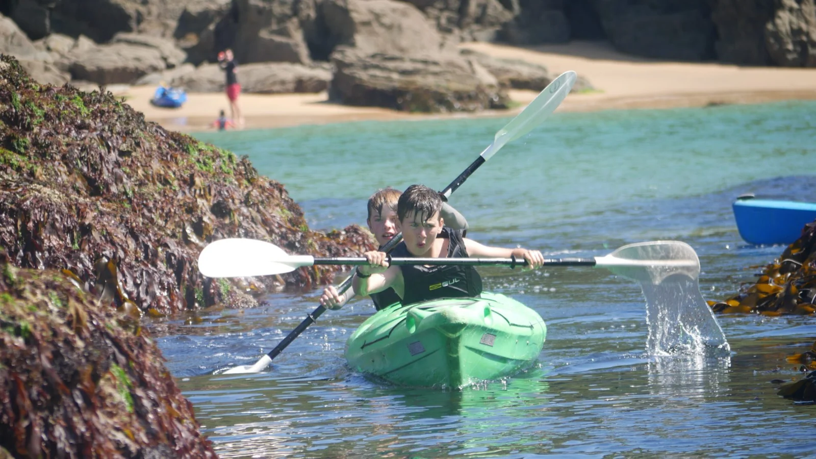 Two paddlers in a green tandem kayak beside coastal rocks