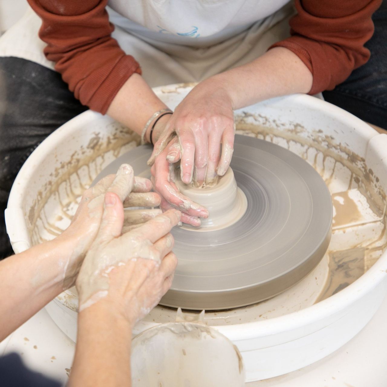 A tutor guiding a visitor’s hands on a pottery wheel