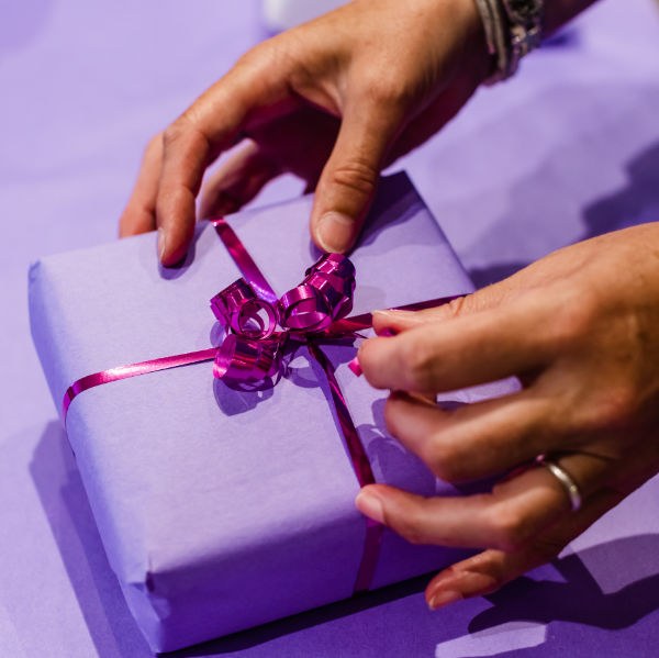Hands tying a pink ribbon around a lilac-wrapped gift at tessdesigns