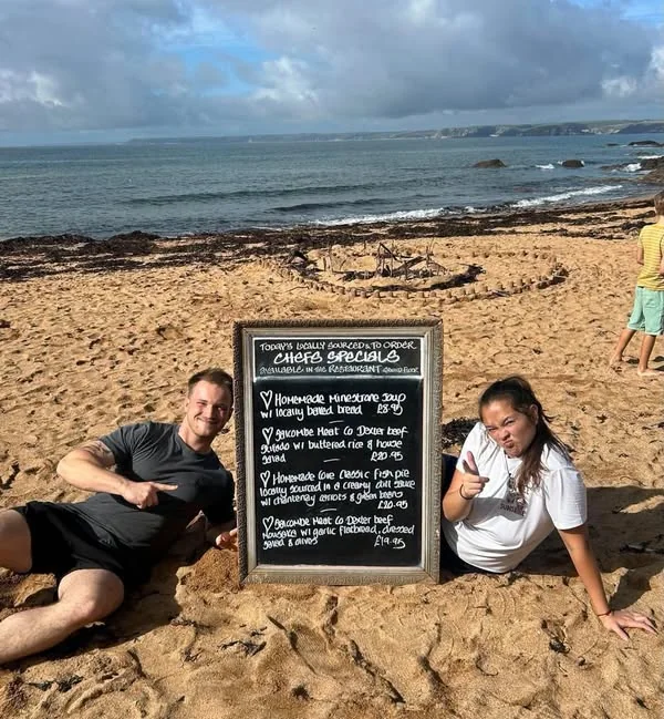 Two members of The Cove team seated beside a specials board on the sand