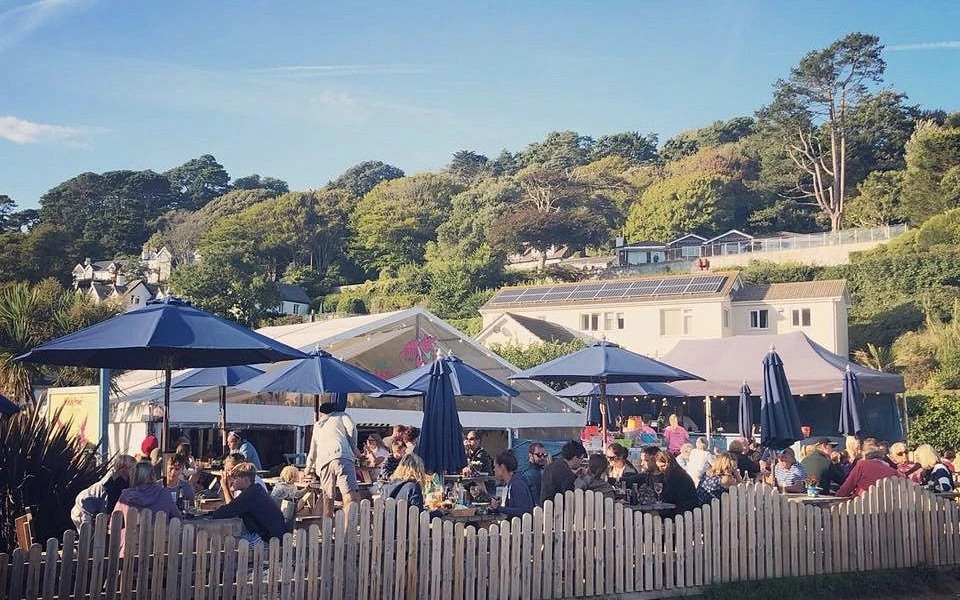 Outdoor tables beneath blue parasols outside The Winking Prawn, with a white picket fence and wooded hillside behind