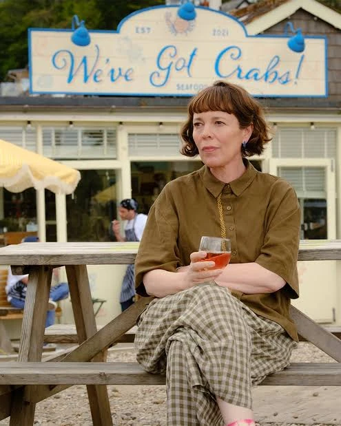 Film still showing a woman seated at a picnic table beneath the fictional We’ve Got Crabs sign